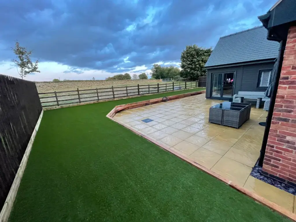 Artificial lawn and sandstone patio installation in Saxilby by landscape gardeners featuring golden paving, raised timber edging and a bright green low maintenance grass area beside a modern grey brick home with countryside views