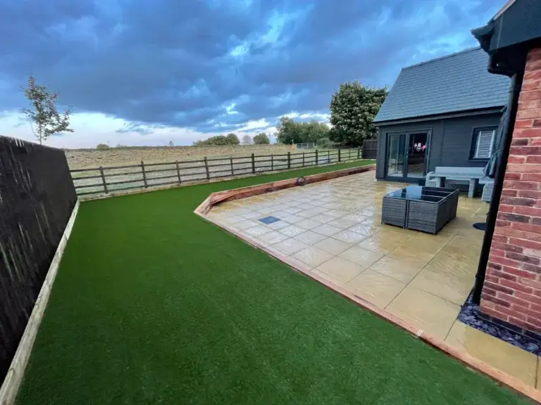 Artificial lawn and sandstone patio installation in Saxilby by landscape gardeners featuring golden paving, raised timber edging and a bright green low maintenance grass area beside a modern grey brick home with countryside views