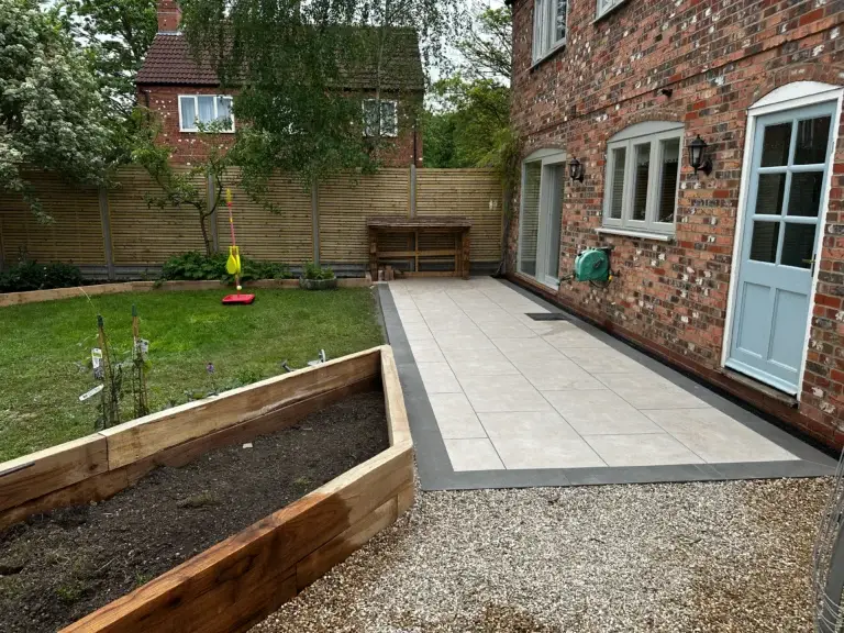 Modern cream porcelain patio bordered with dark grey edging beside a red brick home, featuring raised timber planters and gravel pathways. Installed by Lincoln based landscapers.