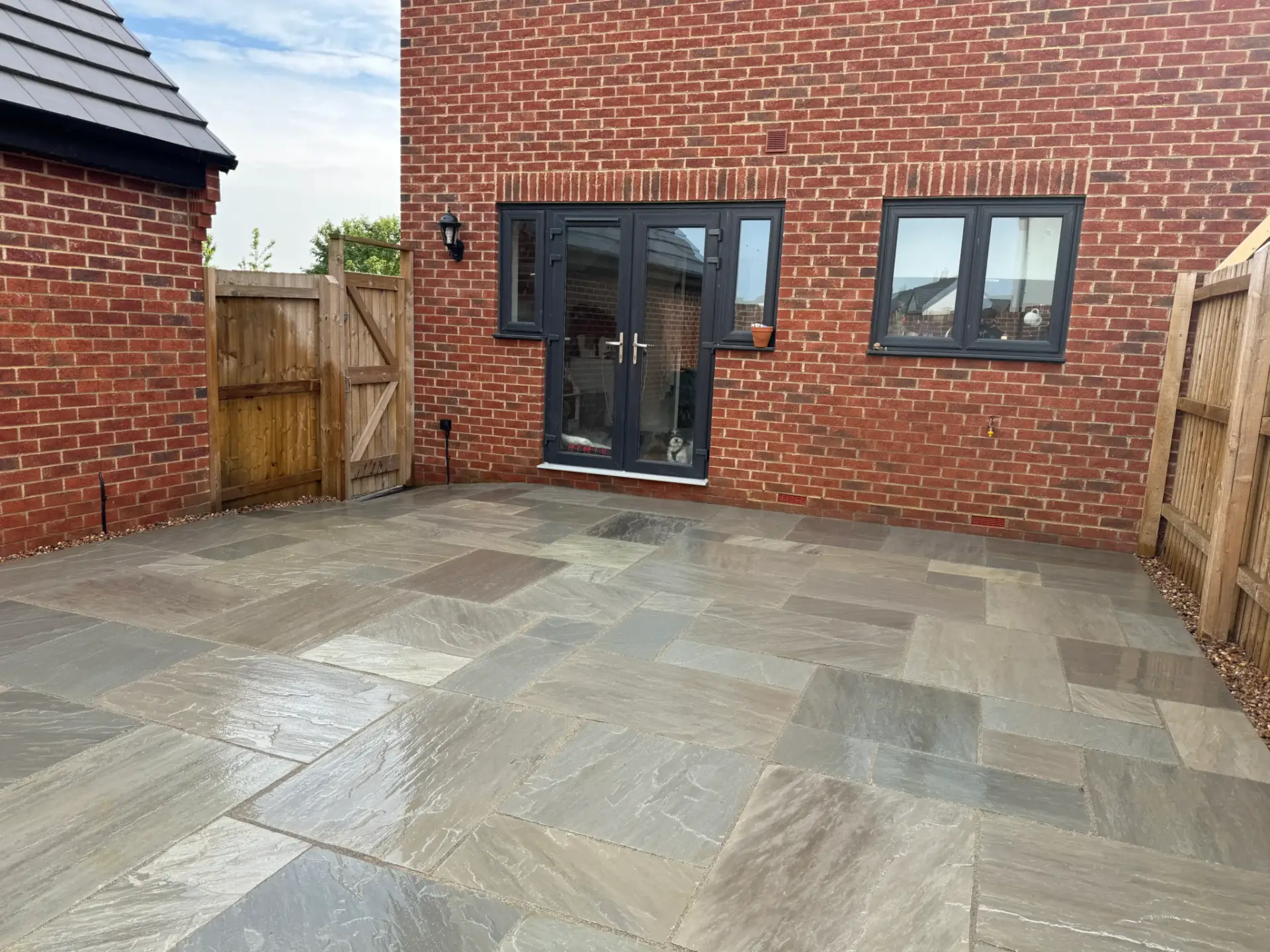 Modern grey sandstone patio installation with a natural textured finish, set against a red brick home and enclosed with timber fencing for a private garden space. Completed by landscapers in Lincoln, Lincolnshire.