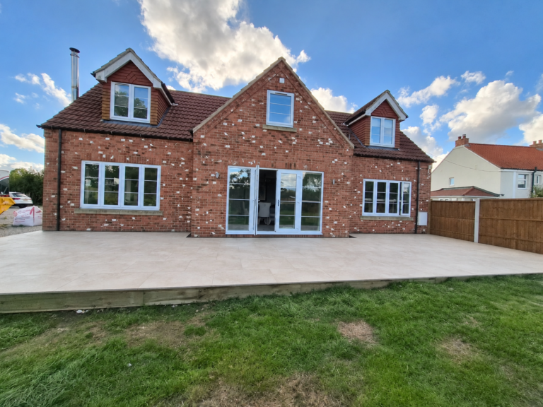 Cream paved terrace spanning the rear of a red-brick house, with timber edging above the garden lawn.