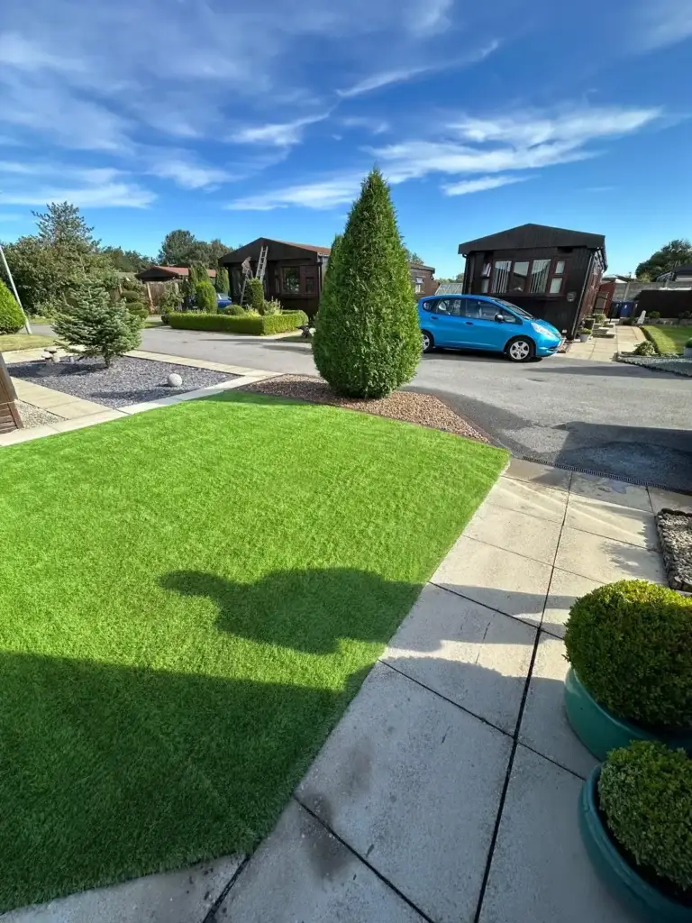 Artificial grass installation at landscape gardeners featuring lush green synthetic turf, cream paving slabs and manicured topiary planters in front of modern wooden lodges under a bright blue sky