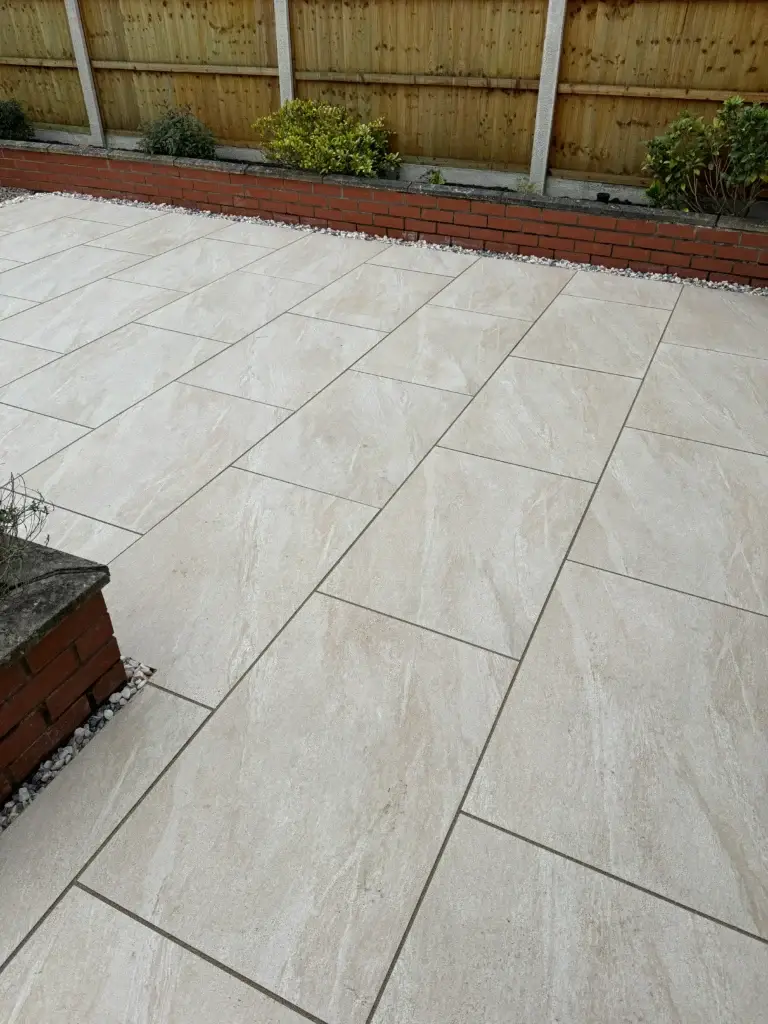 Light cream porcelain patio installation with subtle stone texture and clean joint lines, complemented by low red brick planters and a wooden boundary fence. Completed by landscape gardeners in Gainsborough, Lincolnshire.