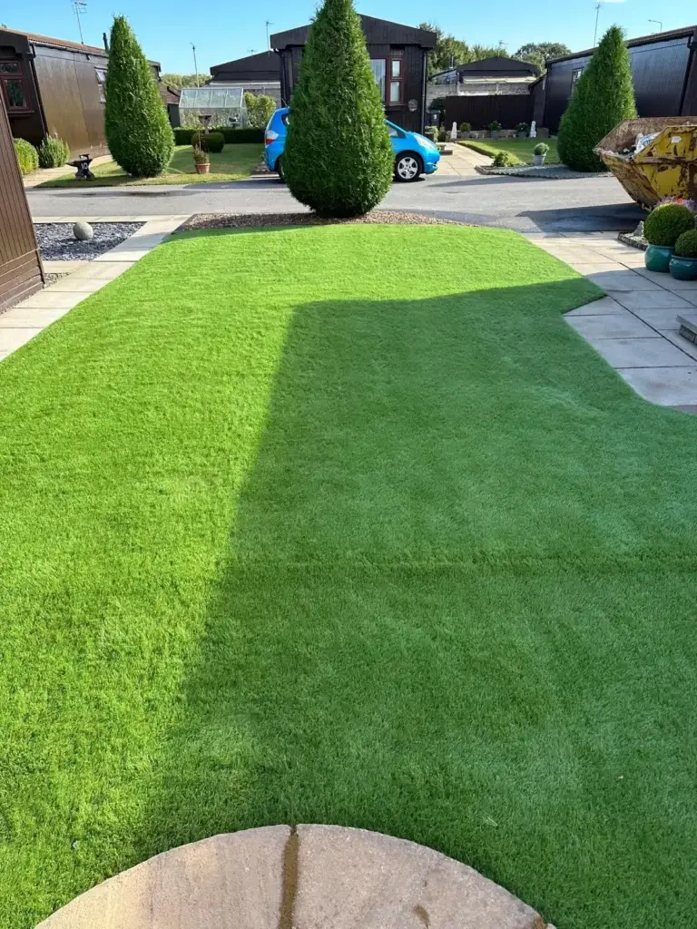 Artificial grass installation and garden design at The Elms Torksey by landscape gardeners featuring bright green synthetic turf framed by cream paving slabs and decorative planting in front of modern wooden lodge homes