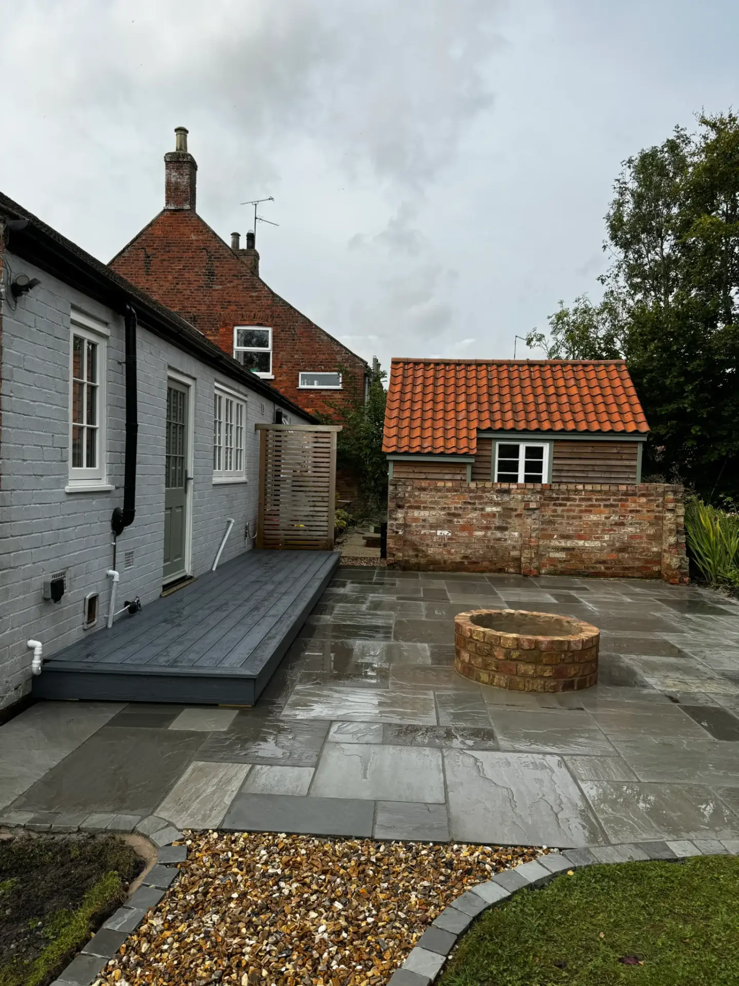 Grey sandstone patio installation with a round brick firepit and raised decking beside a white cottage and tiled garden outbuilding. Completed by garden landscapers in Lincolnshire.