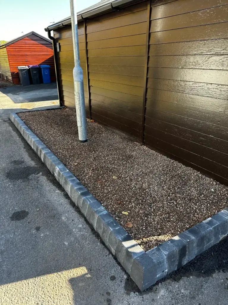 Decorative gravel border installation by landscapers in Lincoln featuring dark brown gravel with clean grey block edging around a street light beside modern wooden lodge exteriors