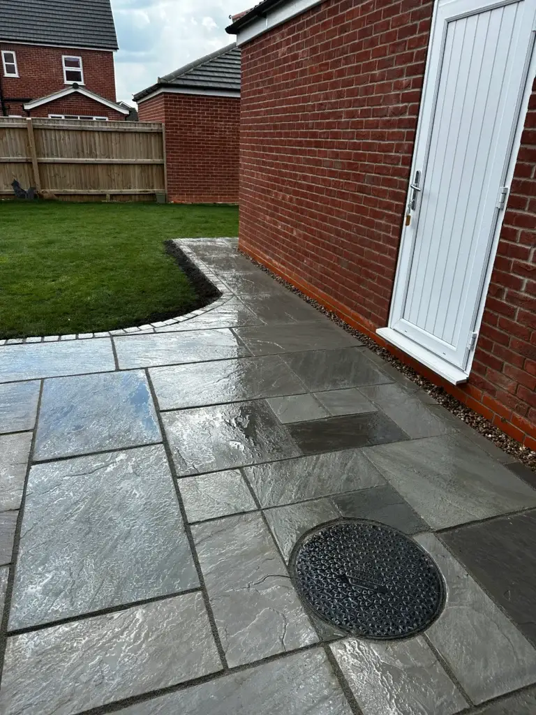 Grey sandstone patio installation with clean-cut edges and natural riven texture alongside a red brick home, leading to a fresh green lawn. Completed by Lincoln based landscapers.