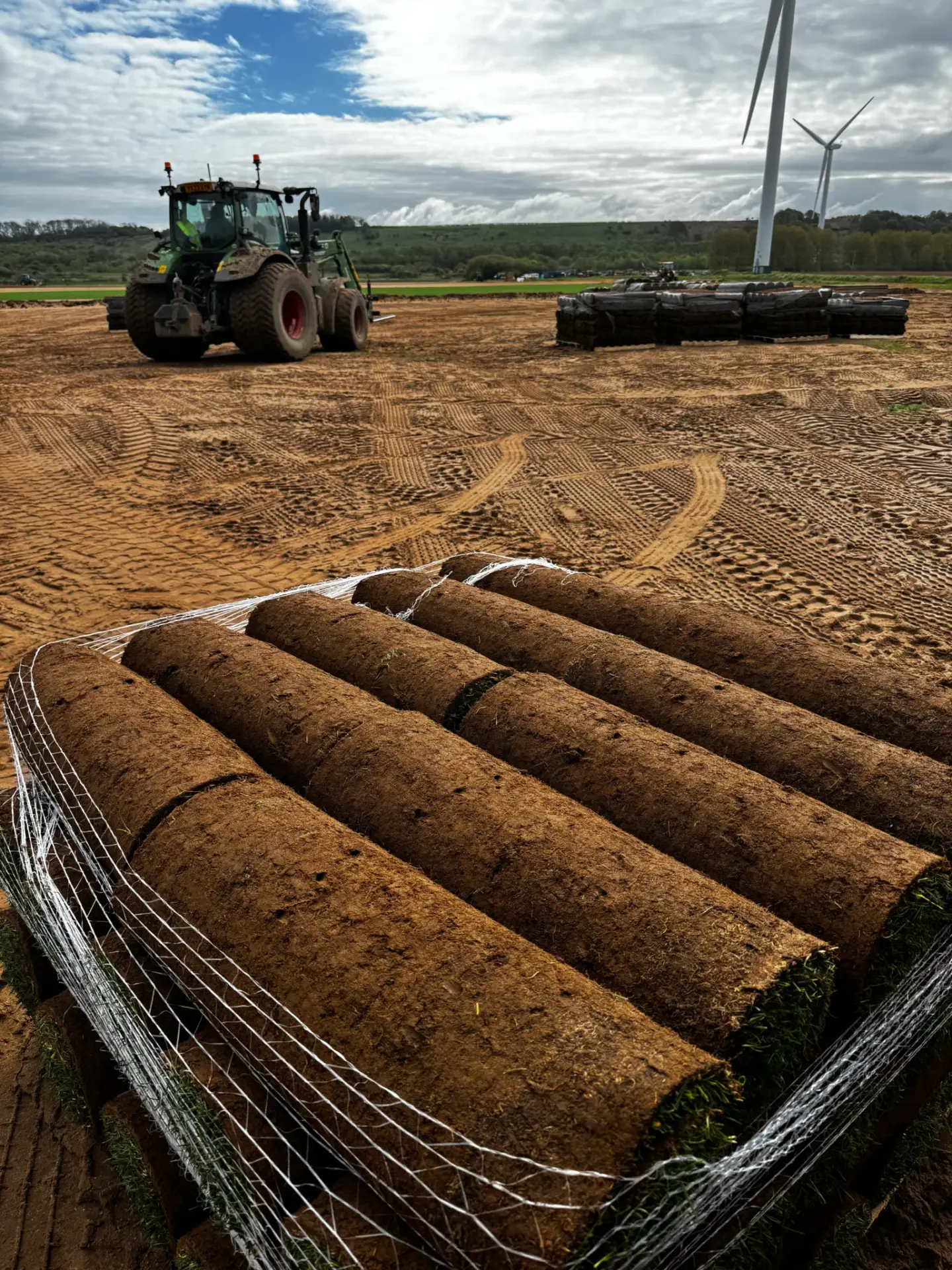 Turf rolls prepared for lawn installation in Lincolnshire by Reshape Landscaping showing stacked rolls of fresh soil-backed grass on a levelled construction site with a tractor and wind turbines in the background