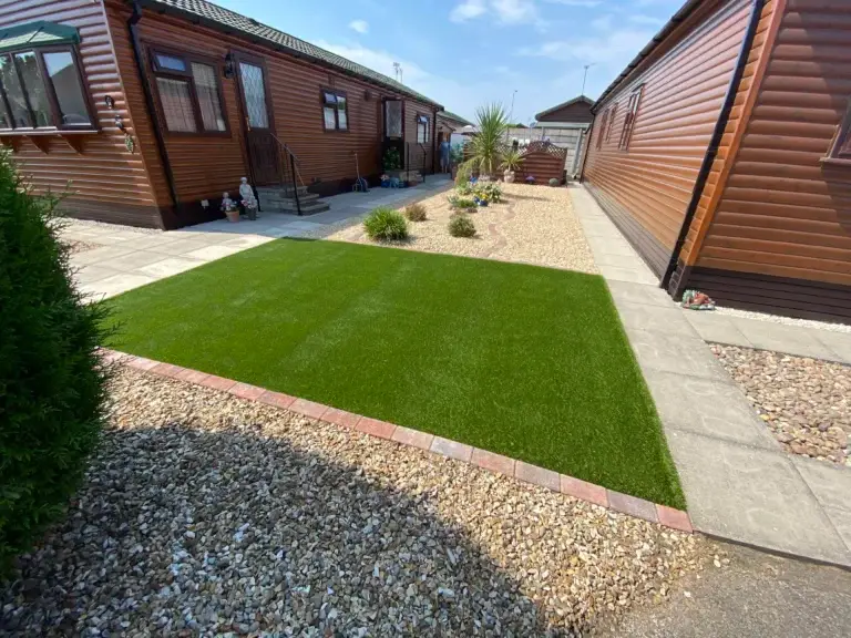 Artificial grass and decorative gravel garden design in Lincoln by landscape gardeners featuring lush green turf bordered with brick edging, golden gravel pathways and modern timber lodges under a clear blue sky
