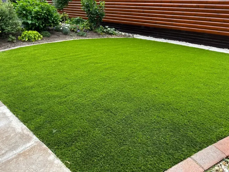 Artificial grass installation at The Elms Torksey by landscape gardeners featuring a deep green low maintenance lawn framed by smooth paving edges and decorative garden borders beside a modern timber lodge