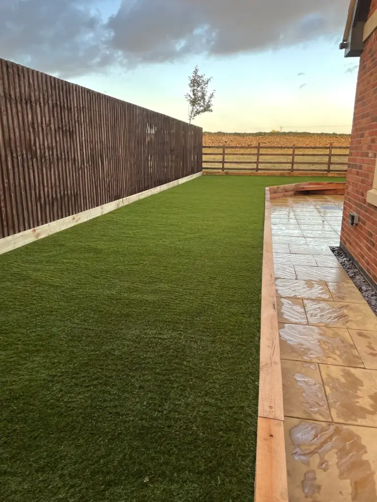 Artificial grass and sandstone patio installation in Saxilby by landscape gardeners featuring golden paving slabs with timber edging and a large low maintenance grass area bordered by wooden fencing behind a modern brick home