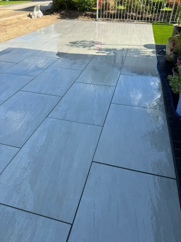 Smooth light grey porcelain patio with precise jointing and a polished wet-look finish, bordered by bright planting and a white garden fence. Installed by Lincoln based landscapers.