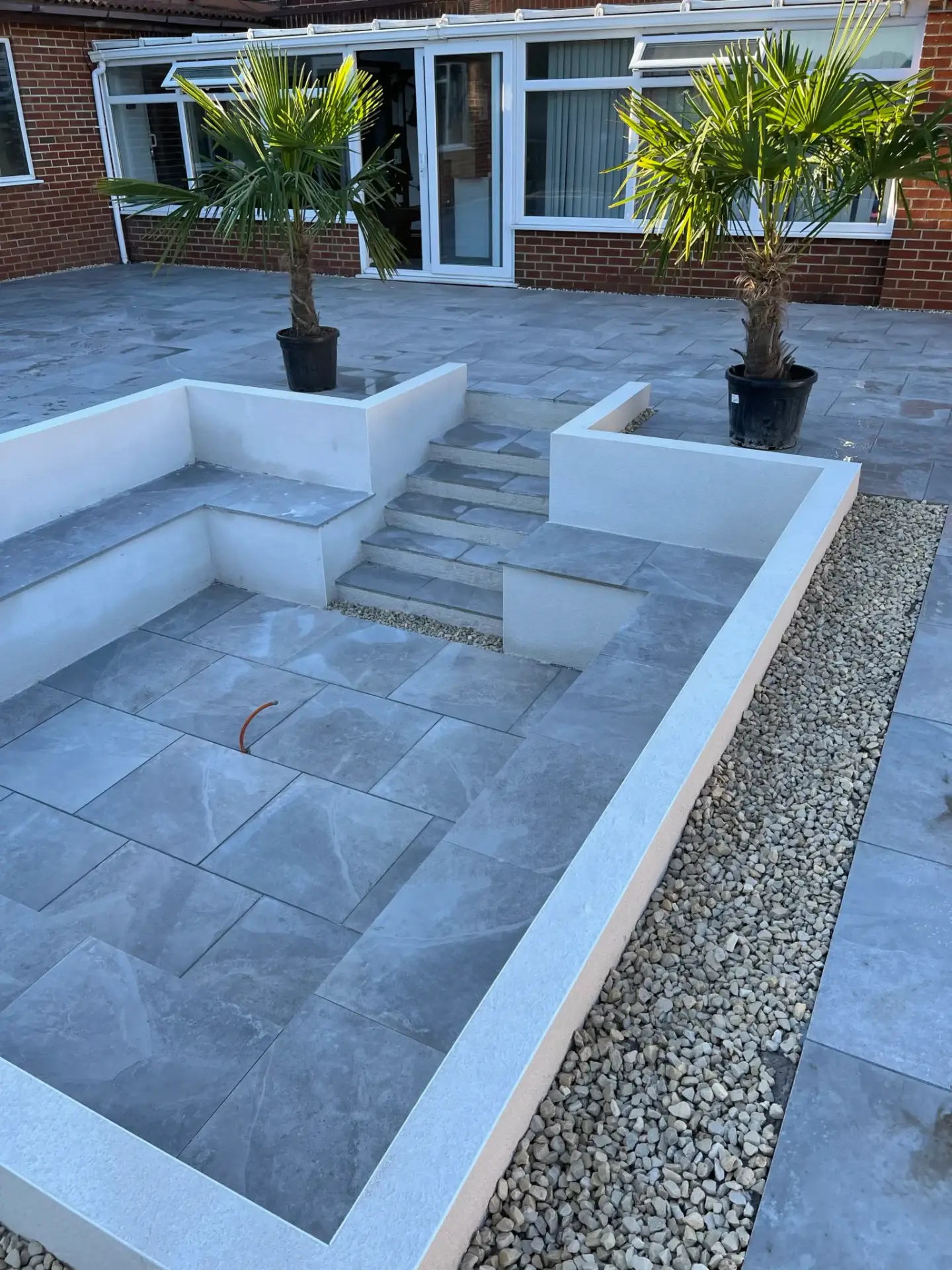 Porcelain patio and sunken seating area installation in Lincoln by landscape gardners featuring smooth grey porcelain tiles, rendered white retaining walls, decorative gravel borders and tropical planting for a luxury outdoor space