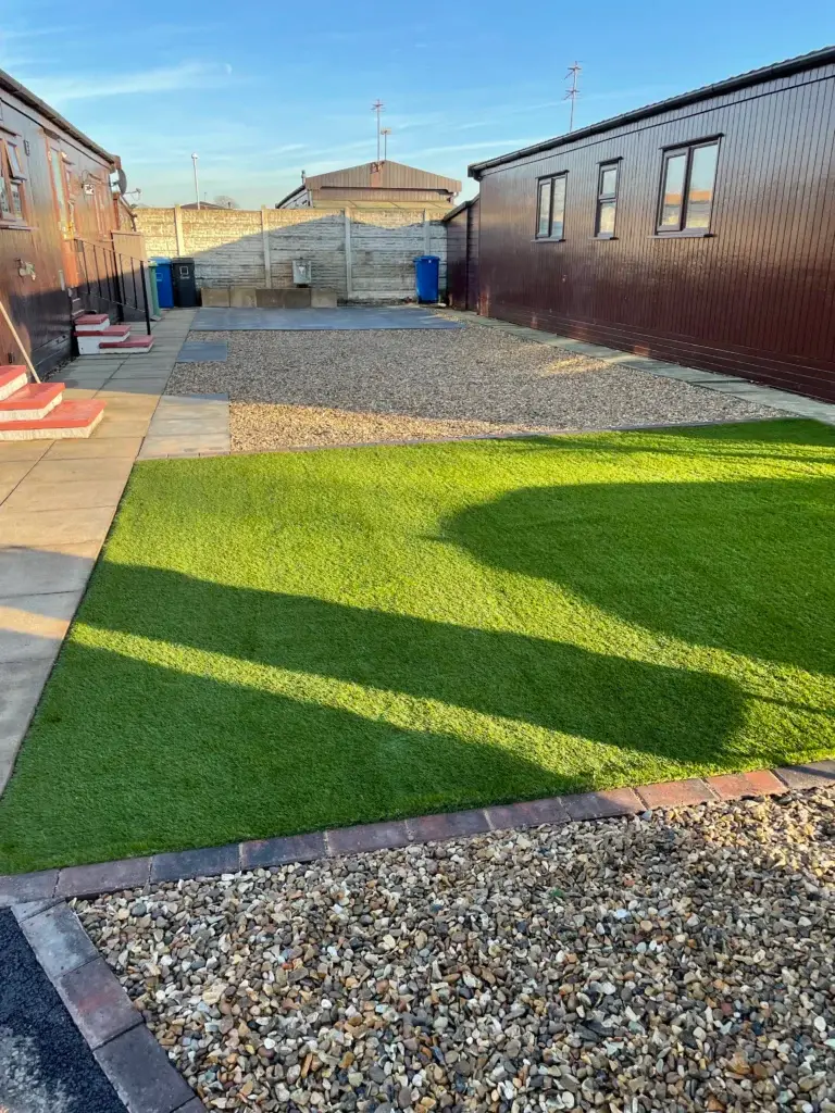 Artificial grass installation and decorative gravel garden design by landscape gardeners featuring green synthetic turf bordered with brick edging, golden gravel areas and modern holiday lodges under a clear blue sky