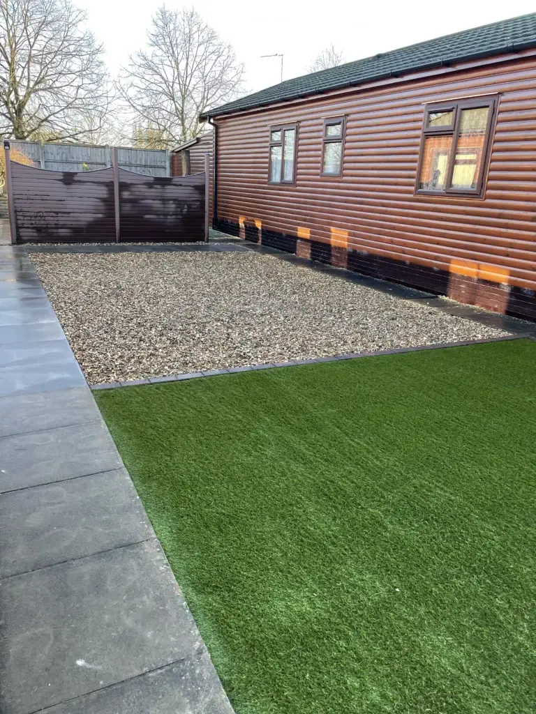 Artificial grass and gravel garden design in Lincoln by landscape gardeners featuring a low maintenance green lawn alongside golden gravel, smooth dark porcelain paving and a modern timber lodge exterior