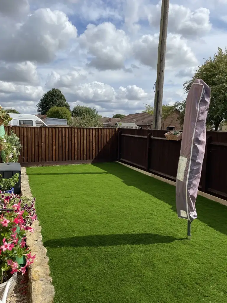 Artificial grass installation and garden design in Lincoln by landscape gardeners featuring a bright green synthetic lawn surrounded by dark timber fencing, stone flower borders and colourful planted flowers under a cloudy sky