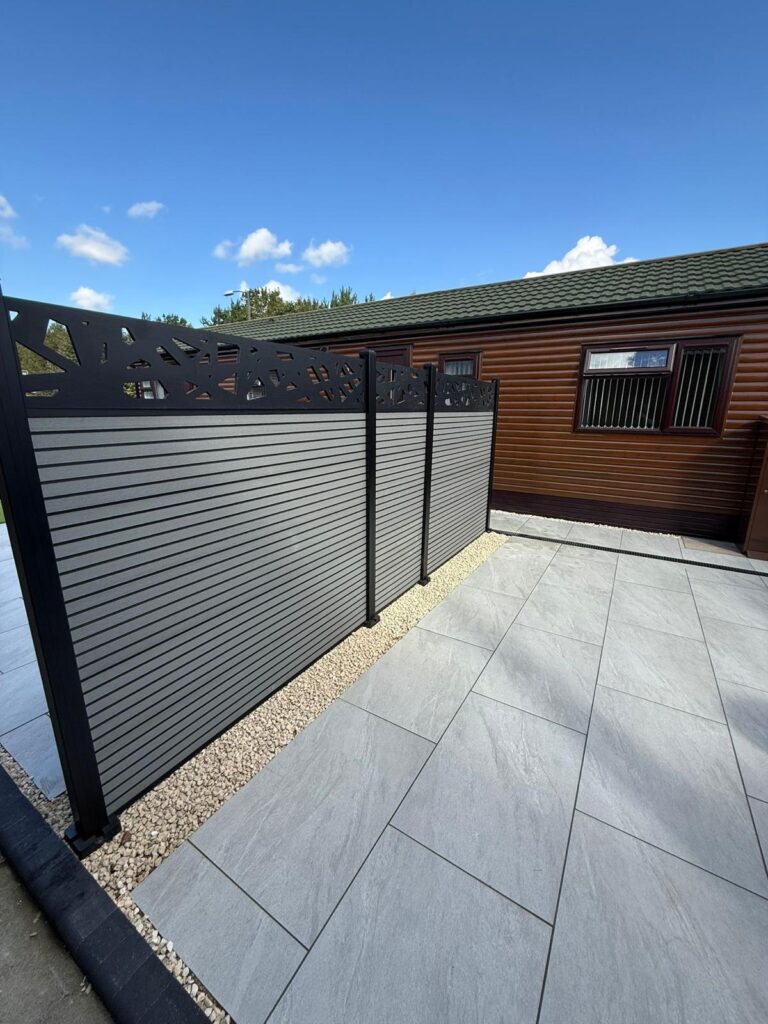 Modern garden patio in Lincoln with large light grey porcelain paving, cream gravel edging, anthracite privacy screening panels and a timber-effect garden lodge backdrop.