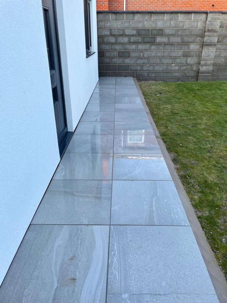 Light grey porcelain side path in Lincoln beside a modern rendered home, with large-format paving, neat lawn edging and dark grey window and door frames.