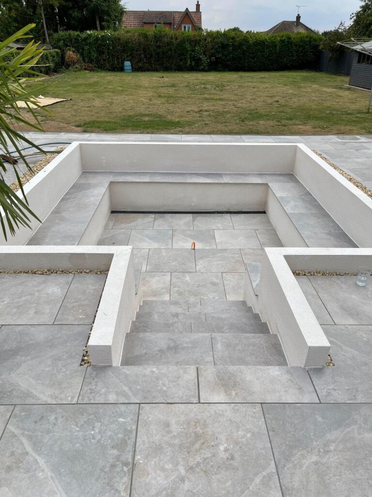 Sunken porcelain patio in Lincoln with light grey large-format paving, rendered seating walls, matching steps and linear drainage channel.