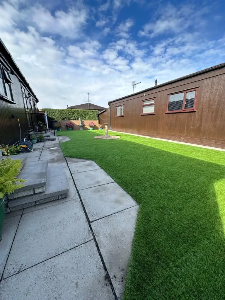 Artificial grass and porcelain paving installation by landscape gardeners featuring bright green synthetic lawn bordered by smooth light grey paving slabs between modern wooden lodges under a blue sky