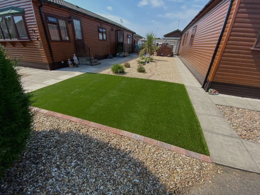 Artificial grass lawn with neat block edging and gravel borders, surrounded by paved walkways and timber lodges for a clean low-maintenance garden. Installed by Reshape Landscaping at The Elms Torksey, Lincolnshire.