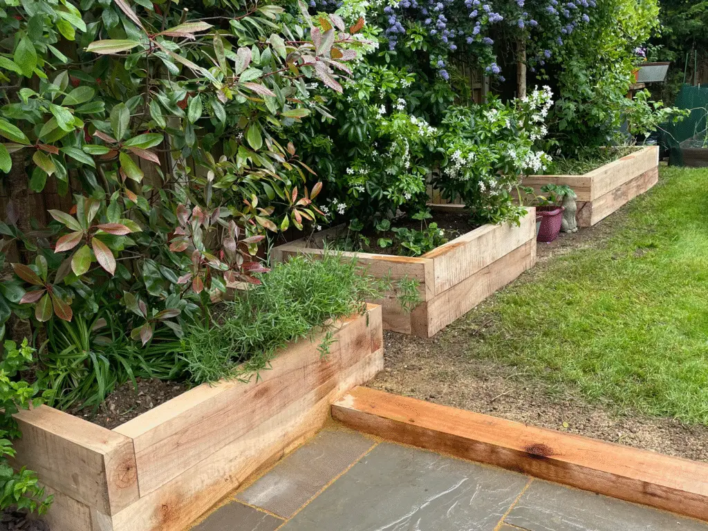 Tiered timber sleeper planters filled with lush green shrubs and flowering plants, bordering a grey sandstone patio for a natural garden design. Built by Reshape Landscaping in Saxilby, Lincolnshire.