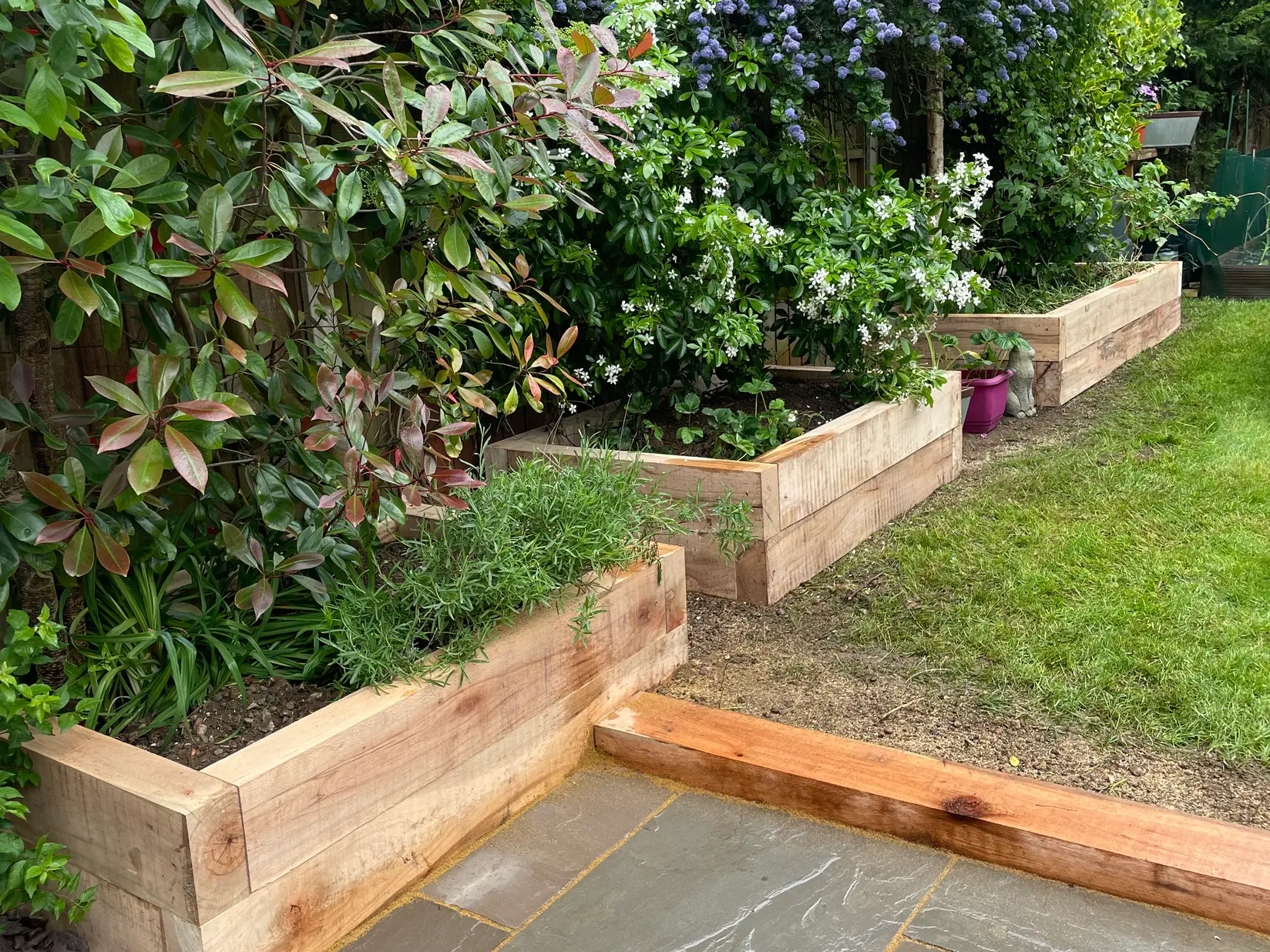 Tiered timber sleeper planters filled with lush green shrubs and flowering plants, bordering a grey sandstone patio for a natural garden design. Built by Reshape Landscaping in Saxilby, Lincolnshire.