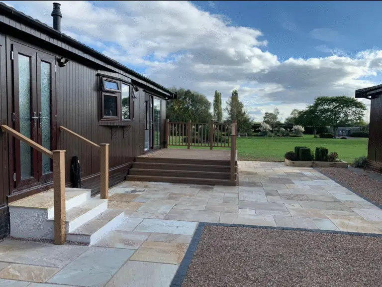 Indian sandstone patio installation in soft buff and cream shades beside a dark timber lodge with raised decking and countryside views. Completed by garden landscapers in Lincolnshire.