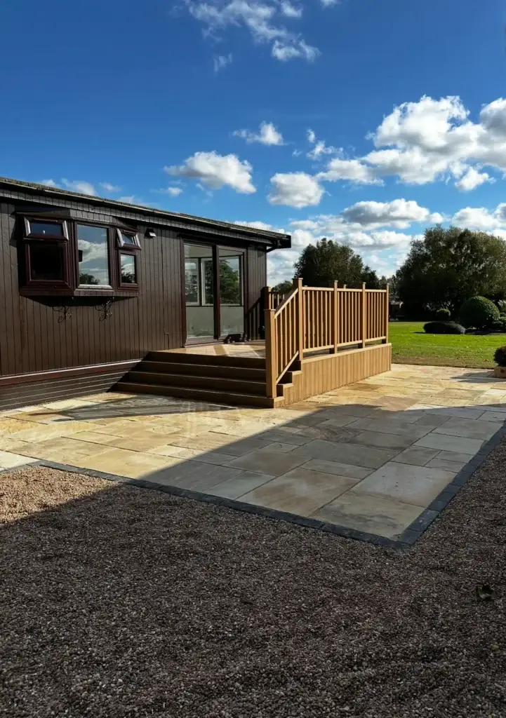 Cream sandstone patio with dark edging and raised composite decking beside a timber lodge, completed landscapers in Lincoln