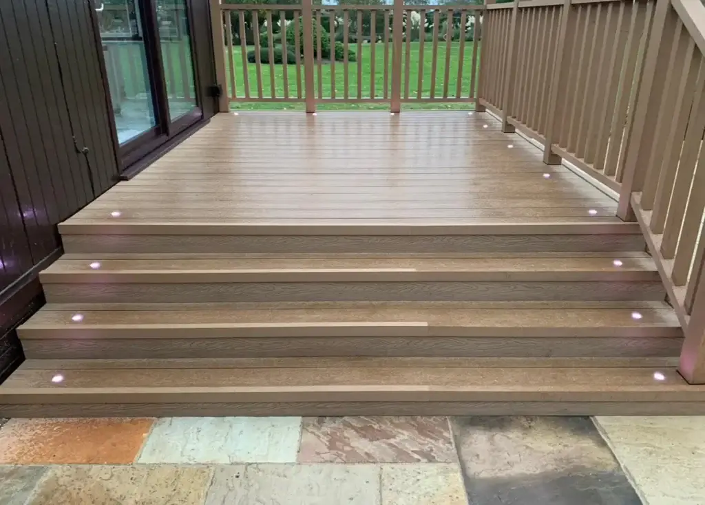 Composite decking installation with warm oak-toned boards and integrated step lighting, built by landscape gardeners at The Elms, Torksey near Lincoln. Modern garden decking with matching handrails overlooking a green lawn