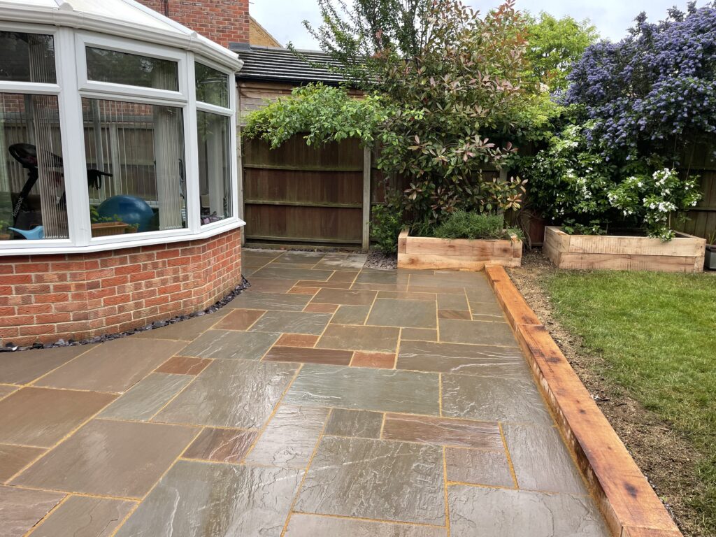 Natural sandstone patio installation in warm brown and grey tones beside a red brick conservatory, finished with raised timber planters and lush green lawn. Completed by landscape gardeners in Gainsborough, Lincolnshire.