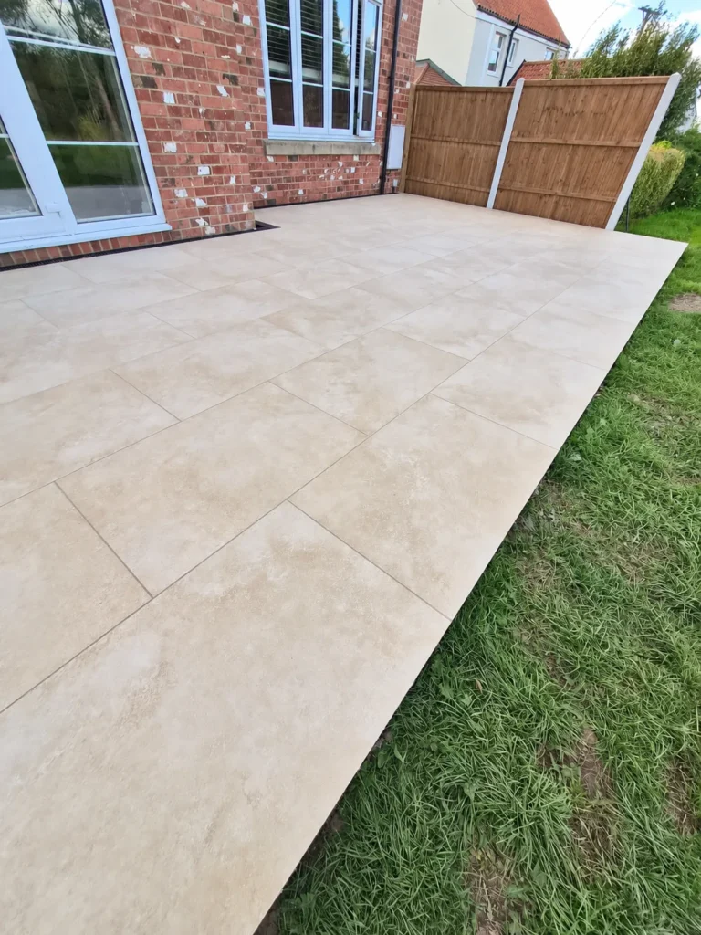 Cream paving slabs forming a patio beside a red-brick house, with a straight lawn edge and timber privacy fencing.