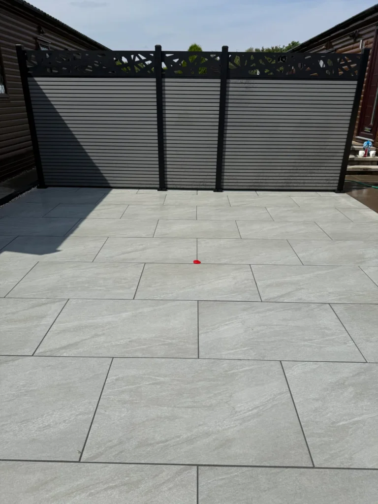 Porcelain patio installation at The Elms Torksey by landscape gardeners in Lincoln featuring large light grey paving slabs with precise joint lines and modern privacy screening between wooden lodge homes for a sleek contemporary finish