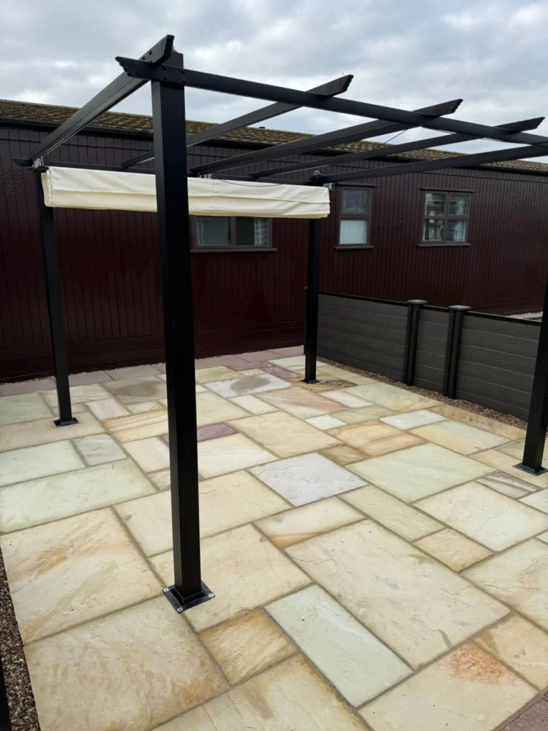 Buff and cream Indian sandstone patio beneath a modern black metal pergola, surrounded by composite fencing and gravel edging for a contemporary garden design. Installed by Lincoln based landscape gardeners.