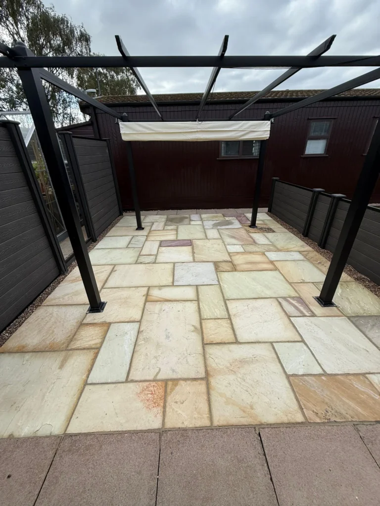Indian sandstone patio in soft buff and cream shades beneath a black metal pergola, finished with smooth jointing and composite fencing for privacy. Installed by Lincoln based landscape gardeners.